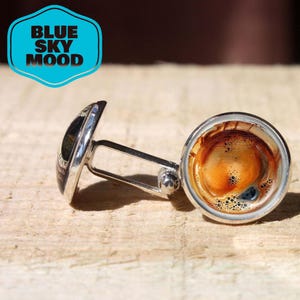 May include: Silver cufflinks with a coffee-themed design. One cufflink features a close-up of coffee, while the other has a black and white design. The cufflinks are on a wooden surface. The text "BLUE SKY MOOD" is in a blue badge.