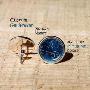 Puede incluir: Un par de pendientes de botón redondos con un fondo azul profundo y arte lineal circular blanco de estilo Gallifreyan. El texto Custom Gallifreyan Words & Names y Available in multiple colors! está impreso cerca de los pendientes.