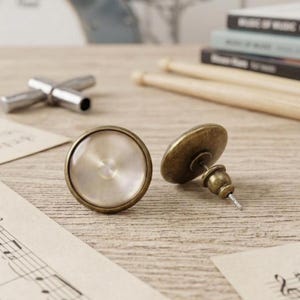 Könnte beinhalten: Ein Paar antike, messingfarbene Ohrstecker mit perlmuttfarbenem, kreisförmigem Design. Die Ohrringe sind auf einer Holzoberfläche platziert, mit einem Stimmschlüssel und Trommelstöcken im Hintergrund. Die Ohrringe haben eine Vintage-Ästhetik.