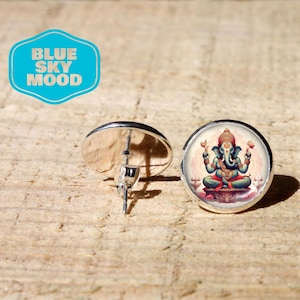 May include: Silver-tone stud earrings featuring a detailed illustration of the Hindu deity Ganesha. The image shows Ganesha in a seated pose, with a teal body, red accents, and a decorative headdress. The earrings are set against a light brown wooden surface. The text "BLUE SKY MOOD" is in a blue bubble.