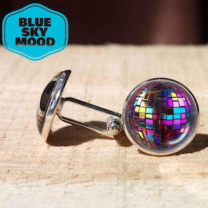 May include: A pair of silver cufflinks with a colorful disco ball design. One cufflink features a black and silver design, while the other showcases a vibrant mosaic of colors. The cufflinks are set against a light brown, textured surface. The image includes the text "BLUE SKY MOOD".