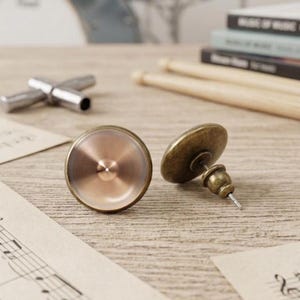 Könnte beinhalten: Ein Paar Ohrstecker mit einem kreisförmigen Design, das wie ein metallisches Schlagzeugbecken aussieht. Die Vorderseite hat ein poliertes kupferfarbenes Finish mit einem Mittelpunkt, während die Rückseite einen dunkelbronzenen Metallstift zeigt.
