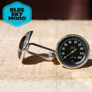 Könnte beinhalten: Silberne Manschettenknöpfe mit schwarzem Tachometerzifferblatt, mit weißen Zahlen und Markierungen. Der Tachometer zeigt bis zu 240 mph an. Die Manschettenknöpfe haben ein klassisches Design und der Text "BLUE SKY MOOD" steht in einem blauen Sechseck.