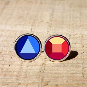 May include: Two round stud earrings with silver-colored borders. One earring features a blue triangle design within a dark blue circle. The other earring has a square design in shades of red and yellow.