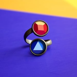 Może przedstawiać: Srebrny pierścionek z dwoma okrągłymi kamieniami szlachetnymi. Górny kamień jest czerwony i ma kształt kwadratu. Dolny kamień jest niebieski i ma kształt trójkąta.
