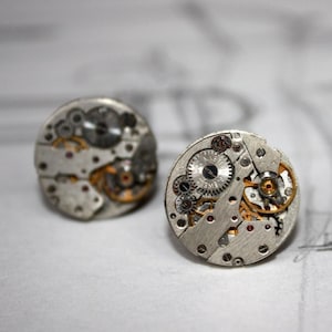 May include: A pair of silver cufflinks featuring exposed watch mechanisms. The intricate internal components, including gears and springs, are visible. These cufflinks have a vintage, industrial style, showcasing the inner workings of a watch.