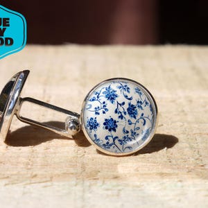 Puede incluir: Gemelos plateados con un diseño floral azul sobre fondo blanco. Los gemelos están sobre una superficie de madera. La imagen incluye un logotipo azul y blanco que dice "BLUE SKY MOOD".