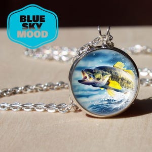 Peut inclure: Un collier pendentif argenté avec un pendentif rond représentant un poisson sautant hors de l'eau. Le poisson a des nageoires jaunes et se détache sur un fond bleu. Le collier a une chaîne et le texte "BLUE SKY MOOD" est visible.