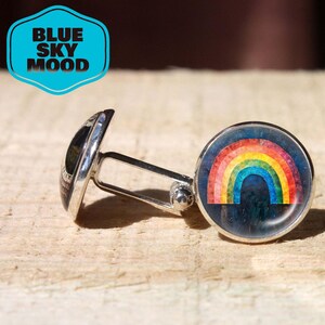 Puede incluir: Gemelos plateados con un diseño de arcoíris colorido. Los gemelos tienen una cara redonda con un gráfico de arcoíris y un respaldo plateado. El texto "BLUE SKY MOOD" está en un gráfico azul en forma de escudo.