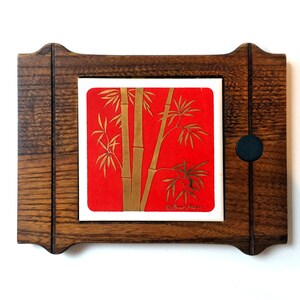 Peut inclure: Un carreau encadré en bois avec un fond rouge et un motif de bambou doré. Le carreau est entouré d'un cadre en bois brun foncé avec un bouton noir sur le côté droit. Le carreau porte le texte "© Fred Press" dans le coin inférieur droit.