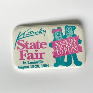 May include: A white button with a pink and green design. It says "Kentucky State Fair" in pink letters and "Your Ticket To Fun!" in pink letters on a white ticket shape. The button also features two cartoon bears holding the ticket.