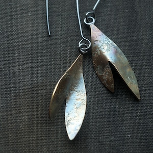 May include: Artisan earrings with a leaf-inspired design. Each earring has a silver wire hook and a bronze-toned metal pendant. The pendants have a textured surface and a split at the base, adding to their unique aesthetic.