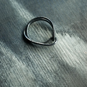 May include: A dark grey metal ring with a textured, uneven surface. The band features an organic, irregular shape that loops over itself, creating a layered appearance with a matte finish and visible metallic grain throughout the entire structure.