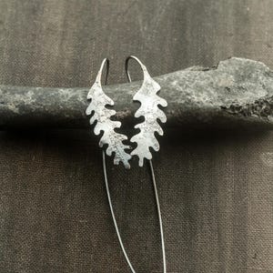 May include: Silver-toned leaf-shaped earrings with a textured surface. The earrings have a long, slender, dangling design. The earrings are displayed on a dark gray stone against a dark gray background.
