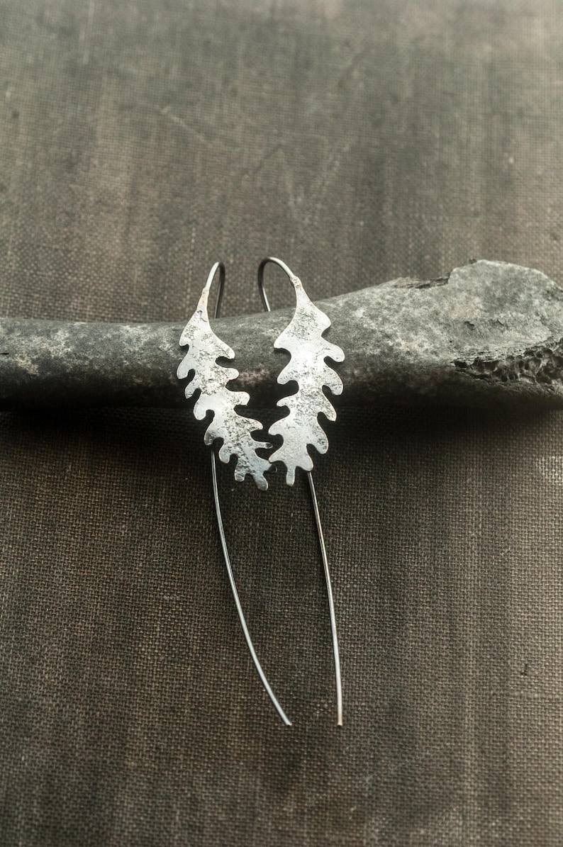 May include: Silver-toned leaf-shaped earrings with a textured surface. The earrings have a long, slender, dangling design. The earrings are displayed on a dark gray stone against a dark gray background.