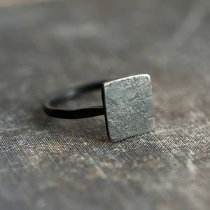 Puede incluir: Un anillo de plata con una parte superior cuadrada y texturizada. El anillo est&aacute; sobre una superficie gris texturizada.