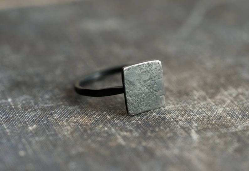 Puede incluir: Un anillo de plata con una parte superior cuadrada y texturizada. El anillo est&aacute; sobre una superficie gris texturizada.