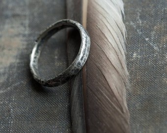 May include: A silver ring with a textured finish, resting on a gray fabric background with a feather beside it.