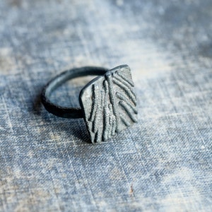 May include: A silver ring with a textured, rectangular design. The ring is made of metal and has a simple band.