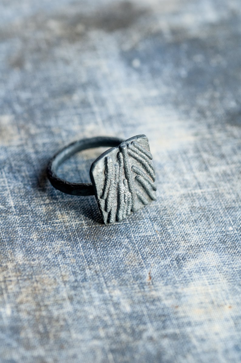 May include: A silver ring with a textured, rectangular design. The ring is made of metal and has a simple band.