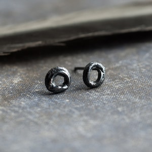 May include: A pair of black oxidized silver earrings with a simple circle design. The earrings are small and delicate, and they have a matte finish.