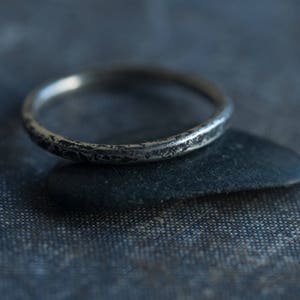 May include: A silver ring with a textured finish. The ring is sitting on a dark gray surface.