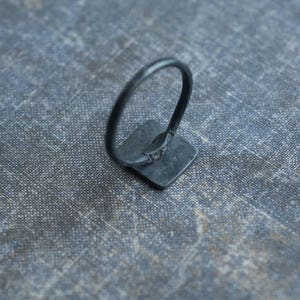 Puede incluir: Un anillo negro con una piedra de forma cuadrada. El anillo es de metal y tiene un dise&ntilde;o sencillo.