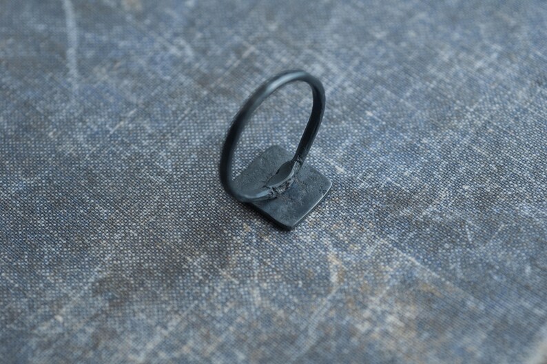Puede incluir: Un anillo negro con una piedra de forma cuadrada. El anillo es de metal y tiene un dise&ntilde;o sencillo.
