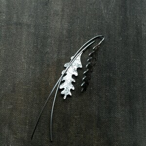 May include: A pair of silver-toned leaf-shaped earrings with a dark, textured background. One earring has a textured, light silver finish, while the other is a solid, dark silver. The earrings have a curved wire design.