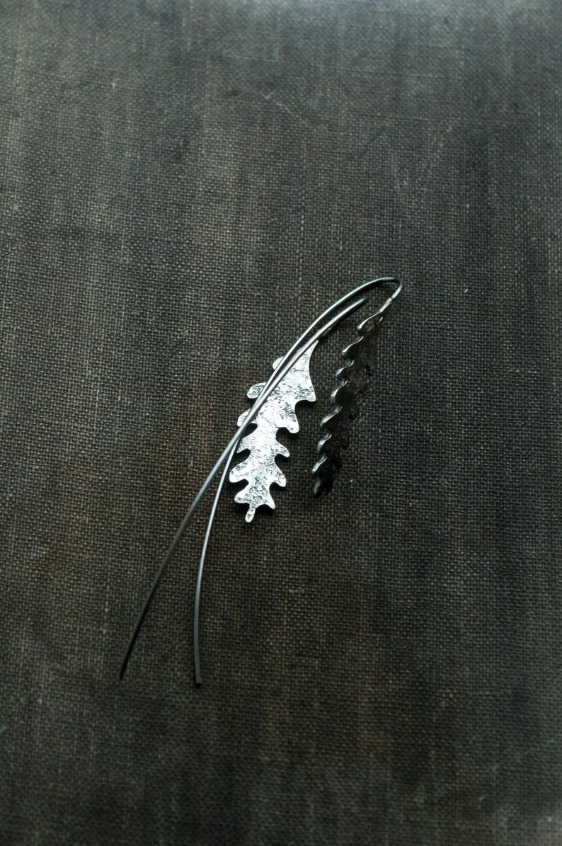 May include: A pair of silver-toned leaf-shaped earrings with a dark, textured background. One earring has a textured, light silver finish, while the other is a solid, dark silver. The earrings have a curved wire design.