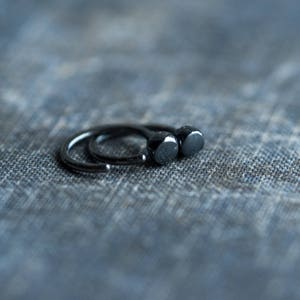 May include: Two black hoop earrings with a rounded, slightly domed shape. The earrings are on a textured blue fabric background.
