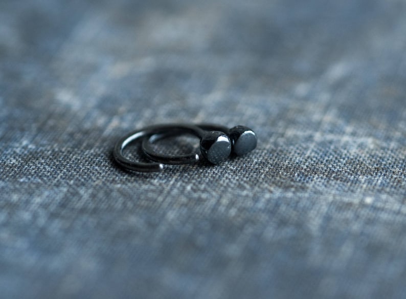 May include: Two black hoop earrings with a rounded, slightly domed shape. The earrings are on a textured blue fabric background.