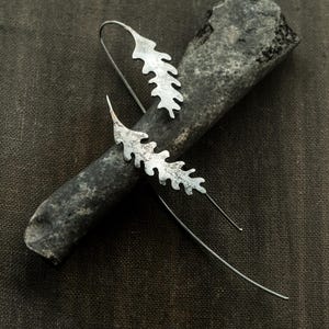 May include: Silver leaf-shaped earrings with long, thin wires. The earrings are displayed on a dark gray, textured surface. The leaf design has a textured, organic appearance, suggesting a nature-inspired jewelry piece.