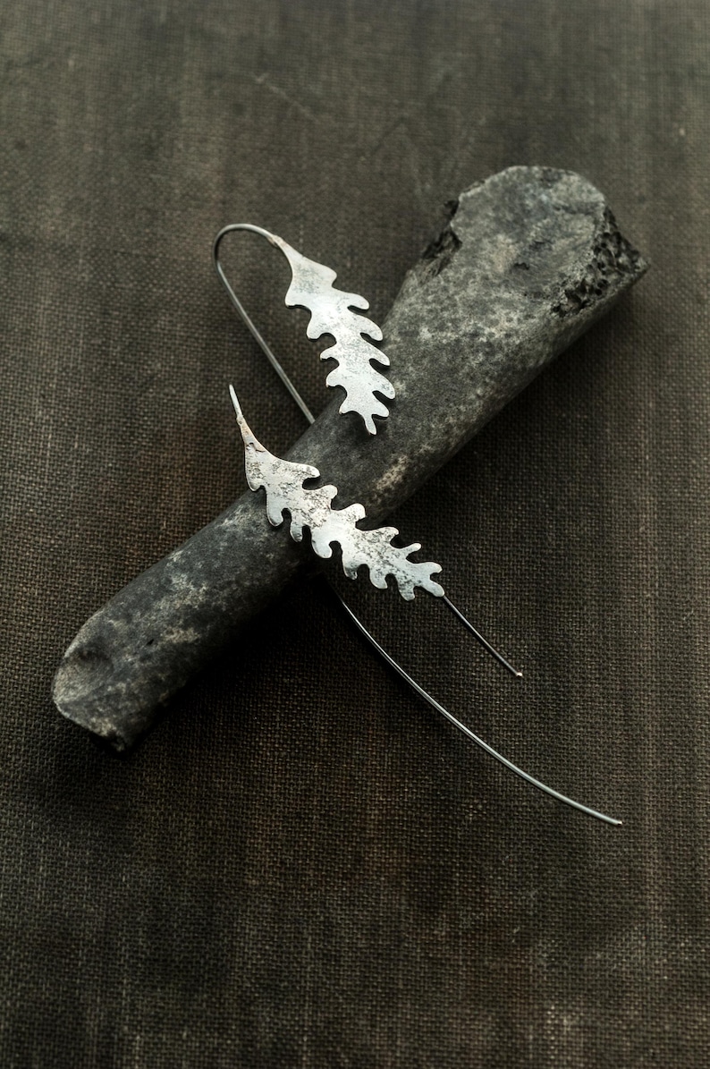 May include: Silver leaf-shaped earrings with long, thin wires. The earrings are displayed on a dark gray, textured surface. The leaf design has a textured, organic appearance, suggesting a nature-inspired jewelry piece.