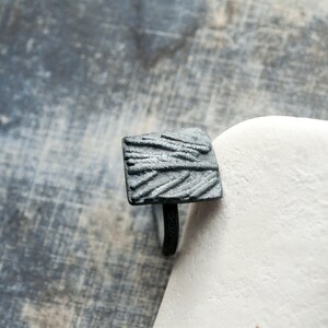 May include: A black square ring with a textured design. The ring is made of metal and has a simple band.