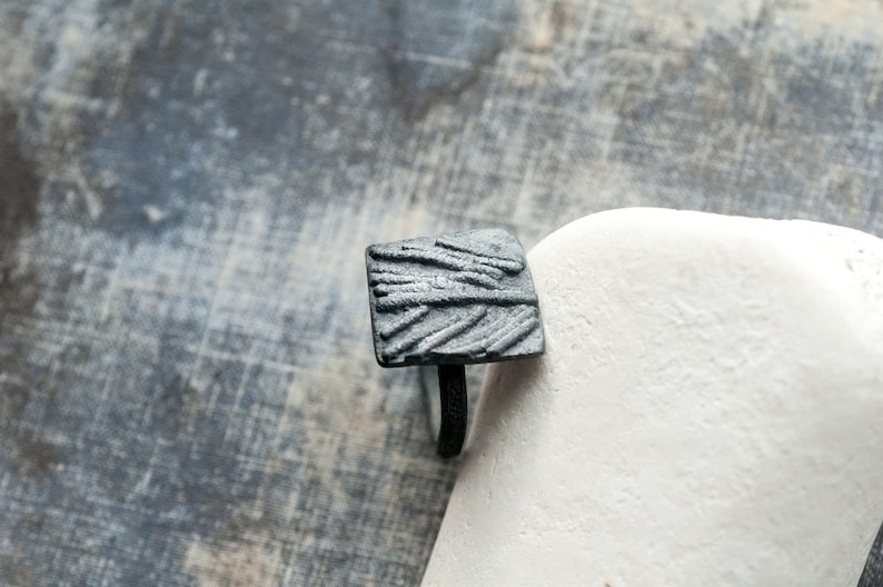 May include: A black square ring with a textured design. The ring is made of metal and has a simple band.