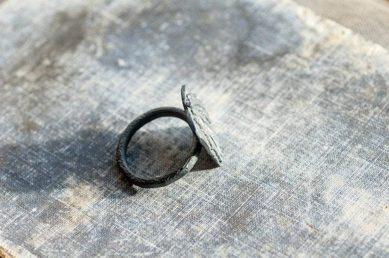 May include: A black metal napkin ring with a decorative design. The ring is lying on a textured surface.