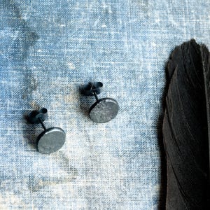 May include: A pair of dark gray stud earrings with a circular, textured face and a post back. The earrings feature a matte, metallic finish with a slightly uneven surface, providing a distinct appearance. Each earring includes a matching metal butterfly clutch.