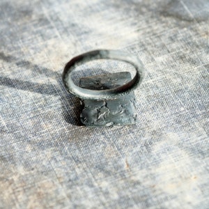 May include: A silver ring with a textured band and a square-shaped setting. The ring is sitting on a textured fabric surface.