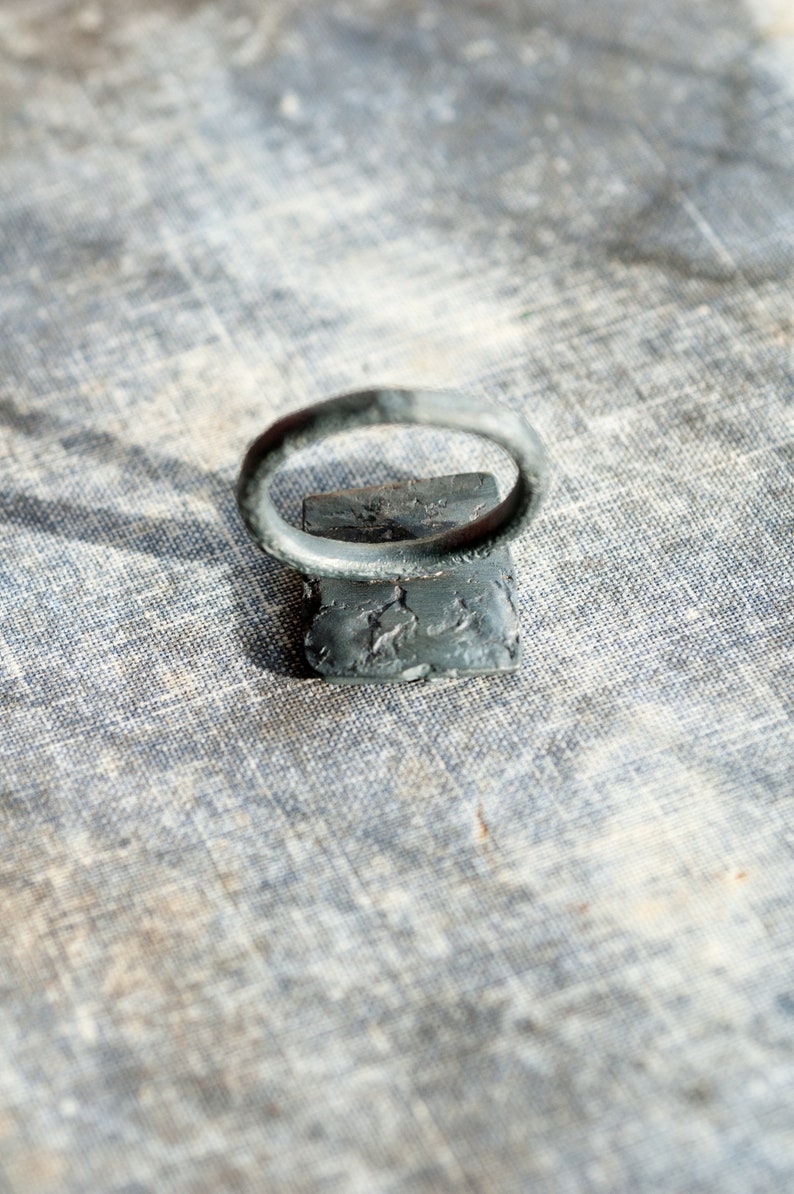 May include: A silver ring with a textured band and a square-shaped setting. The ring is sitting on a textured fabric surface.