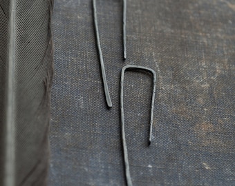 May include: A pair of minimalist, dark gray metal earrings. The earrings have a simple, elongated U-shape design. The background is a textured, dark gray surface with a feather on the left side.