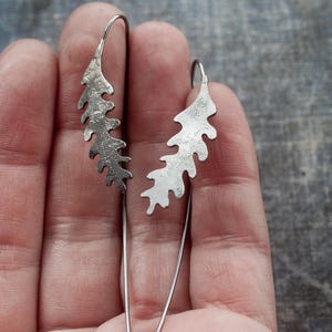 May include: A pair of silver leaf-shaped dangle earrings. Each earring features a textured, stylized leaf design suspended from a thin, curved wire. The earrings are displayed against a neutral background, highlighting their intricate details.