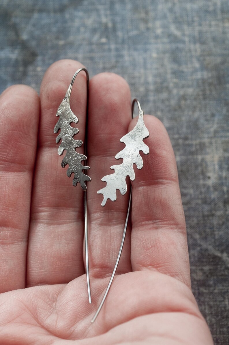 May include: A pair of silver leaf-shaped dangle earrings. Each earring features a textured, stylized leaf design suspended from a thin, curved wire. The earrings are displayed against a neutral background, highlighting their intricate details.