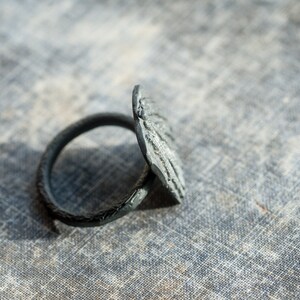 May include: A black metal ring with a textured, circular design. The ring is lying on a textured, gray surface.