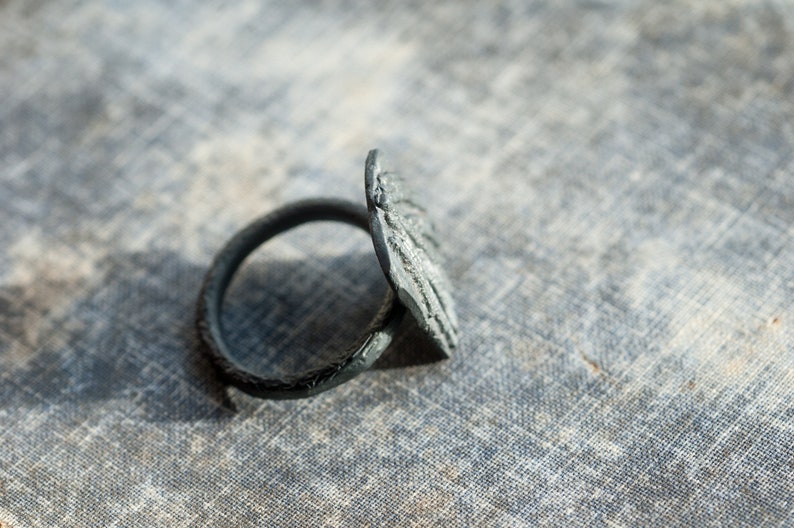 May include: A black metal ring with a textured, circular design. The ring is lying on a textured, gray surface.