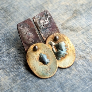 May include: A pair of earrings featuring a rectangular dark copper-toned metal plate attached to an oval-shaped brass-toned metal disc. Each oval disc has a textured surface with a small central metallic bead and a raised, irregular blue-toned accent.