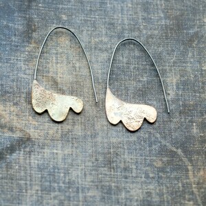 May include: A pair of silver earrings with a textured, organic shape. The earrings are suspended from thin black wires.