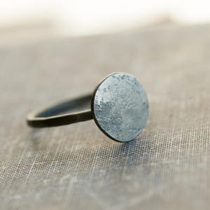 May include: A silver ring with a round, textured top. The ring is made of black metal and has a simple band.