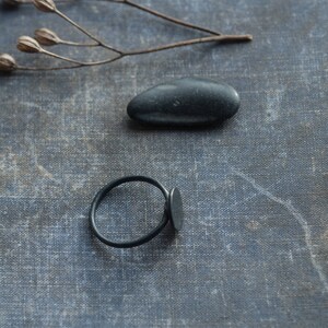 May include: A black ring with a flat oval shaped band and a smooth, black stone. The ring is on a textured, grey surface with a black stone.