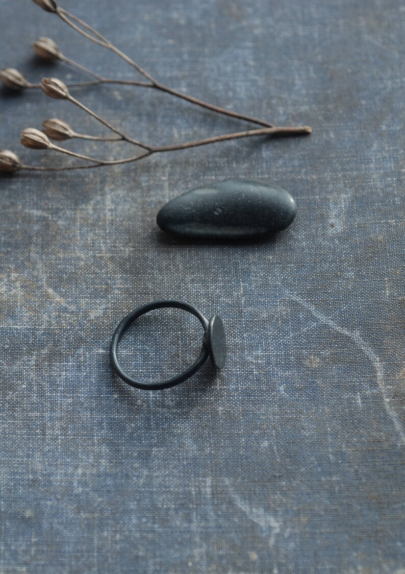 May include: A black ring with a flat oval shaped band and a smooth, black stone. The ring is on a textured, grey surface with a black stone.